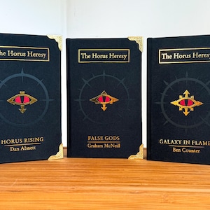 May include: Three hardcover books with the title "The Horus Heresy" in gold lettering. Each book has a black cover with a gold and red eye symbol. The book titles are "Horus Rising", "False Gods", and "Galaxy in Flames".
