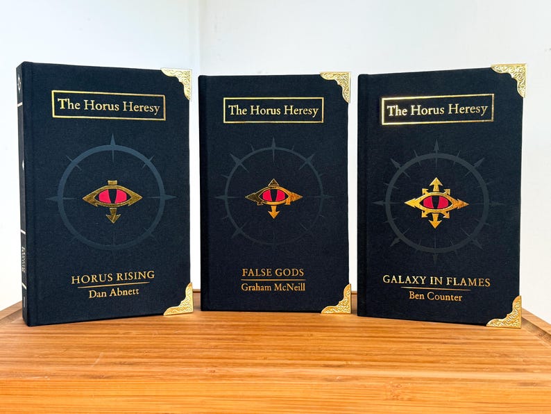 May include: Three hardcover books with the title "The Horus Heresy" in gold lettering. Each book has a black cover with a gold and red eye symbol. The book titles are "Horus Rising", "False Gods", and "Galaxy in Flames".