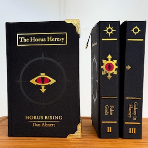 May include: A collection of black hardcover books with gold accents. The first book, "Horus Rising," features the title and author's name in gold, with a red and gold eye symbol. The other two books are labeled "False Gods" and "Galaxy in Flames."