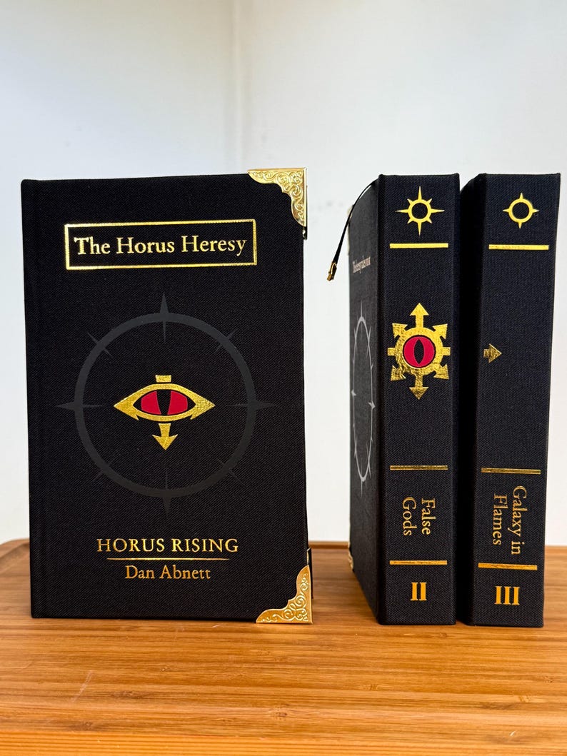 May include: A collection of black hardcover books with gold accents. The first book, "Horus Rising," features the title and author's name in gold, with a red and gold eye symbol. The other two books are labeled "False Gods" and "Galaxy in Flames."