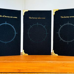 May include: Three black books with gold corner accents and gold lettering. The books have a circular design with a starburst pattern inside. The text on the books reads: "The seeds of heresy are sown", "The heresy takes root", and "The heresy revealed".