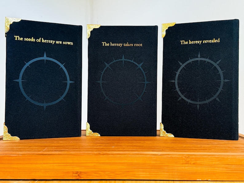 May include: Three black books with gold corner accents and gold lettering. The books have a circular design with a starburst pattern inside. The text on the books reads: "The seeds of heresy are sown", "The heresy takes root", and "The heresy revealed".