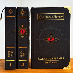 May include: Three hardcover books with black covers and gold lettering. The books are titled "Horus Rising", "False Gods", and "Galaxy in Flames". Each book features a gold emblem with a red eye. The book spines are vertical, and the book covers are horizontal.