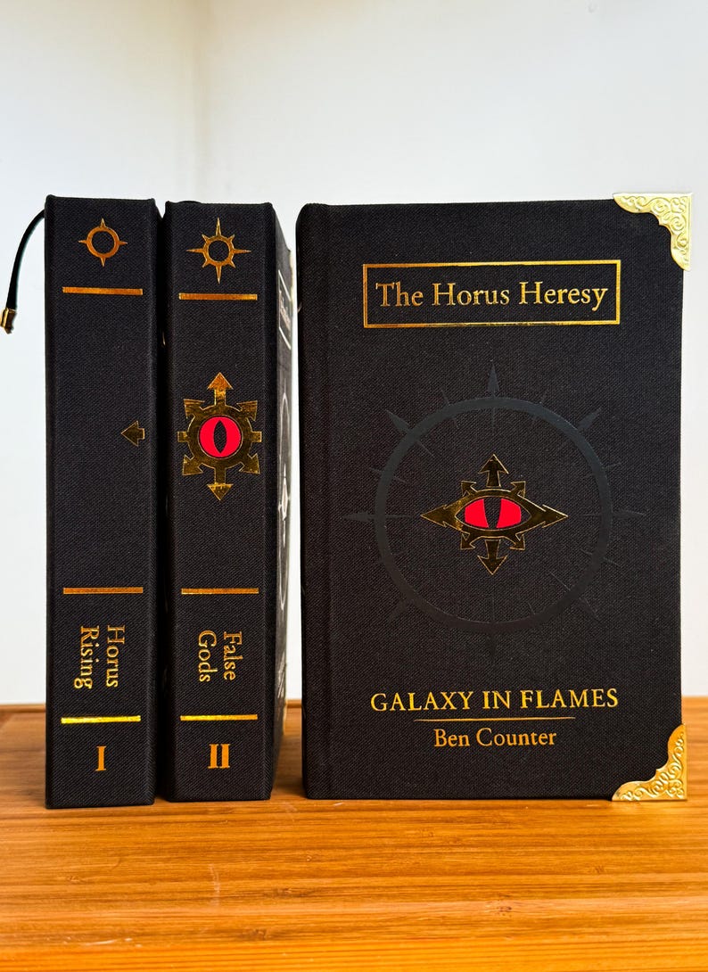 May include: Three hardcover books with black covers and gold lettering. The books are titled "Horus Rising", "False Gods", and "Galaxy in Flames". Each book features a gold emblem with a red eye. The book spines are vertical, and the book covers are horizontal.