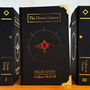 May include: Three hardcover books with black covers and gold lettering. The center book, "The Horus Heresy: False Gods" by Graham McNeill, features a red eye symbol. The other books are labeled "Horus Rising" and "Galaxy in Flames".