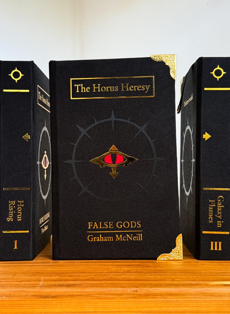 May include: Three hardcover books with black covers and gold lettering. The center book, "The Horus Heresy: False Gods" by Graham McNeill, features a red eye symbol. The other books are labeled "Horus Rising" and "Galaxy in Flames".