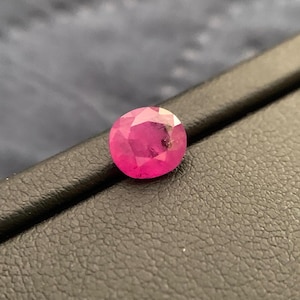 May include: A round, faceted ruby gemstone. The gem is a vibrant shade of pinkish-red, with visible internal inclusions. The ruby is set against a dark, textured surface, highlighting its color and brilliance. This gemstone is suitable for jewelry making.