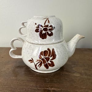 May include: A cream-coloured ceramic teapot and teacup set. The teapot has a rounded body, a curved spout, and a heart-shaped handle. The teacup sits atop the teapot. Both pieces feature brown floral designs and speckled detailing.