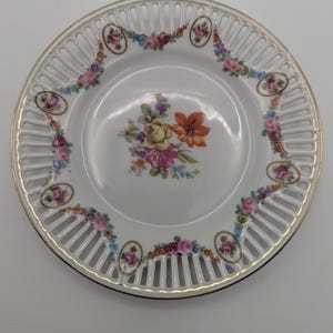 May include: White porcelain plate with a gold rim and intricate openwork design. The plate features a floral bouquet in the center and floral garland around the rim. The flowers are in shades of pink, yellow, and orange.