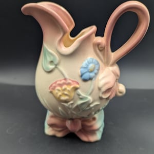 Könnte beinhalten: Vintage Keramikkrug in Pastellfarben. Der Krug hat einen rosa Griff und Rand, mit Blumen- und Schleifendetails in Rosa, Blau und Gelb. Der Sockel wird von einem schleifenförmigen Design getragen.