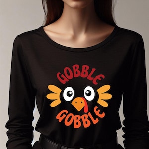 May include: Black long-sleeve shirt with a Thanksgiving-themed graphic. The graphic features a cartoon turkey face with the words "GOBBLE GOBBLE" in red and orange. The turkey has white eyes, an orange beak, and orange feathers.