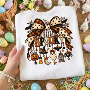 Può includere: Una felpa bianca con una grande grafica di un fiocco decorativo a quadri arancioni e pois color crema. Dal fiocco pendono ciondoli che includono una zucca, un cappello da strega, un fantasma, una lapide con la scritta RIP e un ragno.