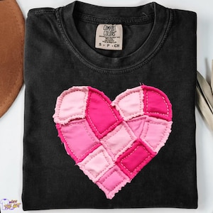 May include: Black t-shirt with a patchwork heart design in shades of pink. The heart is made of various fabric squares stitched together. The shirt has a crew neck and a small tag at the neckline. The shirt is a size small.