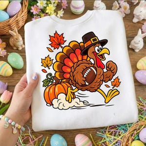 May include: A white sweatshirt featuring a cartoon turkey wearing a pilgrim hat and running while holding a football. The graphic includes an orange pumpkin and several scattered autumn maple leaves in shades of red and orange on the front.