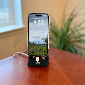 Peut inclure: Un support de téléphone noir avec un câble de charge blanc branché en bas. Un smartphone est dans le support, affichant une image paysage d'un champ vert avec un ciel nuageux. L'heure sur l'écran du téléphone est 4:29. Le support est sur une surface en bois.