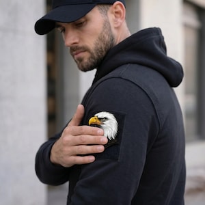 May include: Black hoodie with an eagle head patch. The patch shows a detailed eagle with white feathers and a yellow beak. The patch is rectangular and attached to the upper arm of the hoodie.