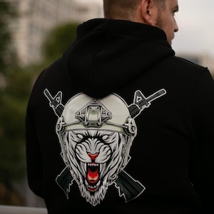 Op de afbeelding: Zwarte hoodie met een afbeelding van een witte tijger met een militaire helm en gekruiste geweren. De tijger heeft een rode neus en een open mond. De hoodie heeft een capuchon en is gemaakt van een zacht materiaal.