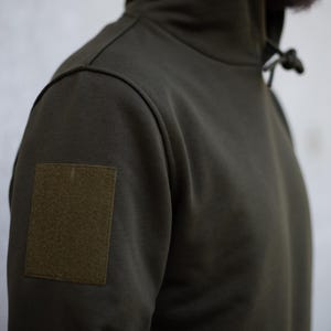 Puede incluir: Sudadera con capucha verde oliva con un parche en la manga izquierda. La sudadera tiene una capucha con cordón y un ajuste relajado. La tela parece ser un material de peso medio, adecuado para climas más fríos.