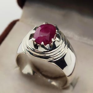 Op de afbeelding: Een zilveren ring met een grote, ovaal geslepen robijn edelsteen. De ring heeft een gepolijste afwerking en een gedetailleerde zetting die de rode edelsteen stevig vasthoudt. De ring wordt gepresenteerd in een sieradendoos.