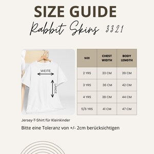 Peut inclure: Un guide des tailles pour les t-shirts Rabbit Skins 3321. Le guide indique les mesures de la largeur de la poitrine et de la longueur du corps en centim&egrave;tres pour les tailles 2 ans &agrave; 5/6 ans. Un t-shirt blanc est affich&eacute;.