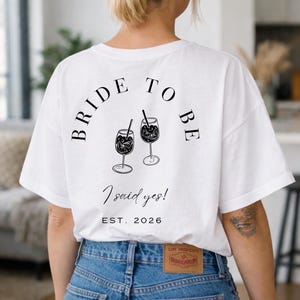 May include: White t-shirt with the text "BRIDE TO BE" arched above two cocktail glasses with straws. Below the glasses, the text reads "I said yes!" and "EST. 2026". The person is wearing blue jeans.