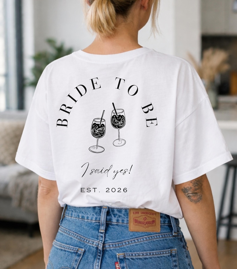 May include: White t-shirt with the text "BRIDE TO BE" arched above two cocktail glasses with straws. Below the glasses, the text reads "I said yes!" and "EST. 2026". The person is wearing blue jeans.