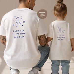 May include: White t-shirts with matching designs. The shirts feature a blue crescent moon and star graphic, with the text "I LOVE YOU TO THE MOON AND BACK." The image shows a parent and child wearing the shirts, promoting a partner look.