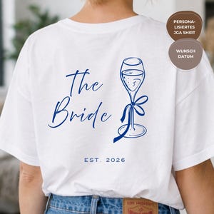 Op de afbeelding: Wit T-shirt met een blauwe afbeelding van een champagneglas met een strik. De woorden "The Bride" zijn in blauw geschreven, met "EST. 2026" eronder. Een bruin label is zichtbaar op de jeans.