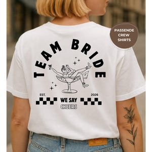 Puede incluir: Camiseta blanca con la frase "TEAM BRIDE" en negro en forma de arco. Debajo, una ilustraci&oacute;n de una mujer en una copa de champ&aacute;n. La camiseta tambi&eacute;n tiene el texto "EST. WE SAY CHEERS 2026".