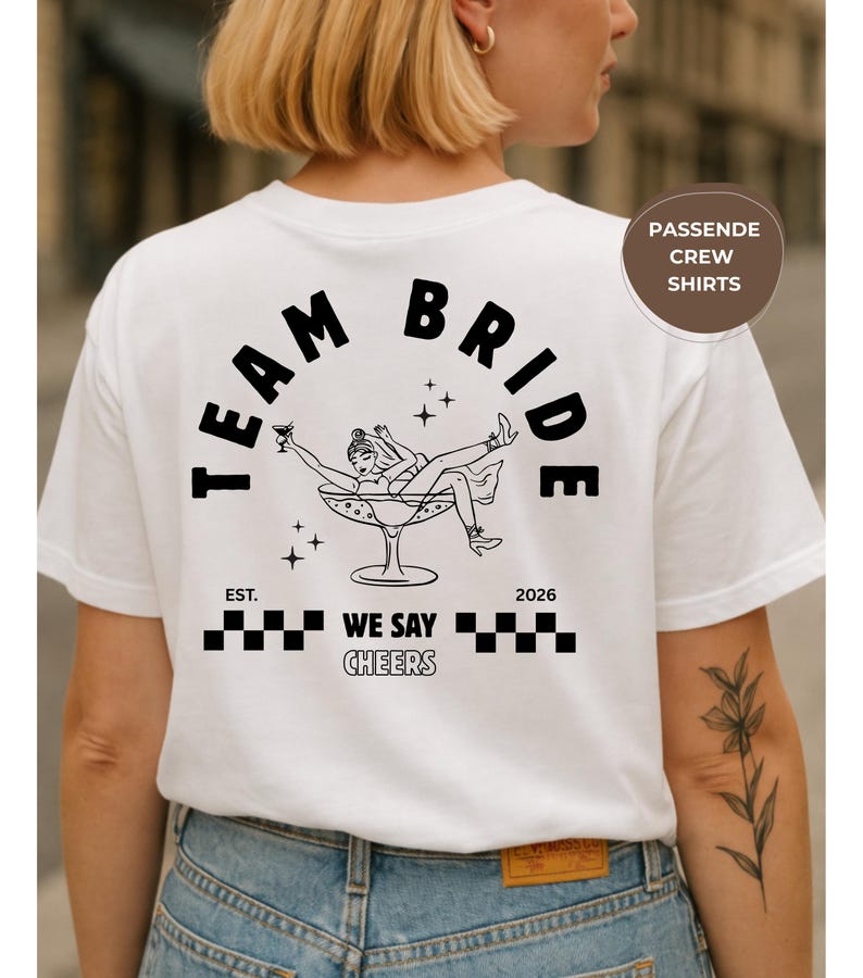 Puede incluir: Camiseta blanca con la frase "TEAM BRIDE" en negro en forma de arco. Debajo, una ilustraci&oacute;n de una mujer en una copa de champ&aacute;n. La camiseta tambi&eacute;n tiene el texto "EST. WE SAY CHEERS 2026".