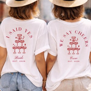 May include: Two white t-shirts with red text and graphics. One says "I SAID YES Bride EST. 2026" and the other says "WE SAY CHEERS Crew EST. 2026". Both have a champagne glass pyramid design, perfect for a hen party.