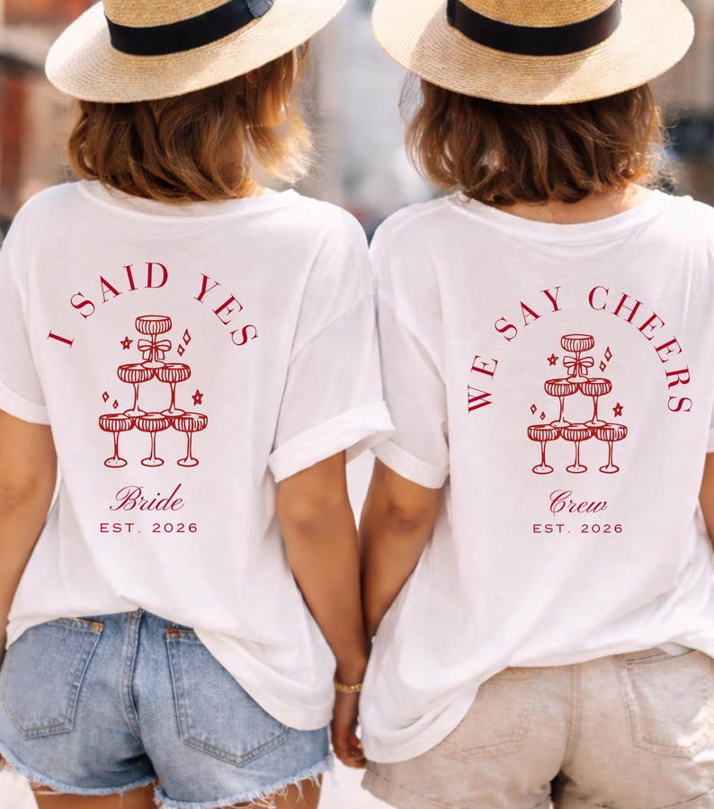 May include: Two white t-shirts with red text and graphics. One says "I SAID YES Bride EST. 2026" and the other says "WE SAY CHEERS Crew EST. 2026". Both have a champagne glass pyramid design, perfect for a hen party.