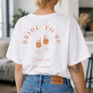 May include: White t-shirt with the text "BRIDE TO BE" in a curved design, along with the phrase "I said yes!" and the year "EST. 2026". The shirt features an illustration of two cocktails. The model is wearing blue jeans.