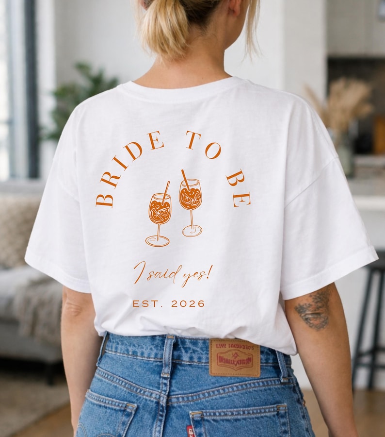 May include: White t-shirt with the text "BRIDE TO BE" in a curved design, along with the phrase "I said yes!" and the year "EST. 2026". The shirt features an illustration of two cocktails. The model is wearing blue jeans.