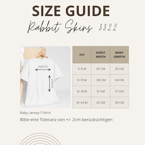 May include: A size guide for Rabbit Skins baby jersey t-shirts. The guide shows chest width and body length measurements in centimeters for sizes 3-6 months to 18-24 months. The text also includes the phrase "Bitte eine Toleranz von +/- 2cm ber&uuml;cksichtigen."