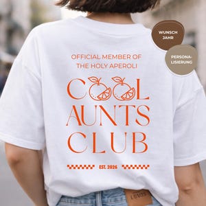Puede incluir: Camiseta blanca con texto naranja « COOL AUNTS CLUB » con gráficos de cítricos naranjas. El texto « OFFICIAL MEMBER OF THE HOLY APEROLI » está encima del texto principal. La camiseta también tiene « EST. 2026 » impreso.