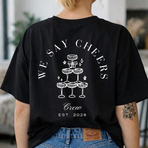 May include: Black t-shirt with a white graphic on the back. The design shows champagne glasses stacked in a pyramid, with the words "WE SAY CHEERS" and "Crew EST. 2026" arranged in a circle.