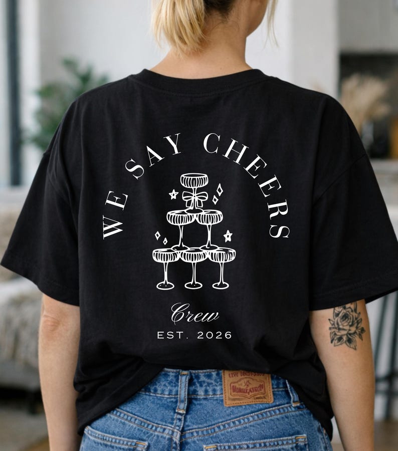 May include: Black t-shirt with a white graphic on the back. The design shows champagne glasses stacked in a pyramid, with the words "WE SAY CHEERS" and "Crew EST. 2026" arranged in a circle.