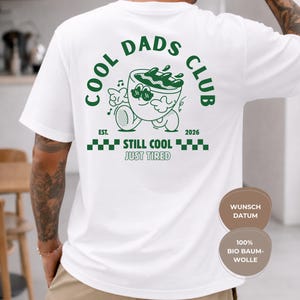 Könnte beinhalten: Weißes T-Shirt mit grünem Aufdruck auf dem Rücken. Der Aufdruck zeigt "COOL DADS CLUB" mit einem Cartoon-Kaffeebecher-Design. Darunter stehen die Worte "EST. 2026" und "STILL COOL JUST TIRED".
