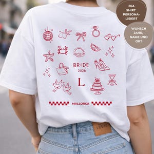 May include: White t-shirt featuring red illustrations of various objects, such as a ring, high heels, and a cake. The word "BRIDE" and the year "2026" are printed in red. The word "MALLORCA" is also printed on the shirt.