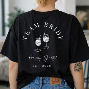 May include: Black t-shirt with the words "TEAM BRIDE" arched above two illustrated spritz cocktails. Below, the text reads "We say Spritz!" and "EST. 2026". The shirt is a casual style.