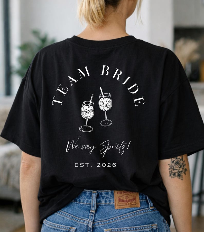 May include: Black t-shirt with the words "TEAM BRIDE" arched above two illustrated spritz cocktails. Below, the text reads "We say Spritz!" and "EST. 2026". The shirt is a casual style.