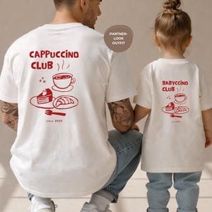 May include: White t-shirts with red graphics. One shirt reads "Cappuccino Club" with coffee cup, cake, and croissant illustrations. The other shirt says "Babyccino Club" with similar graphics. Both shirts have "since 2023" text.
