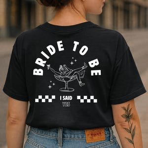 Puede incluir: Camiseta negra con un gr&aacute;fico blanco que dice "BRIDE TO BE" en arco. El gr&aacute;fico presenta a una mujer en una copa de champ&aacute;n, con el texto "I SAID YES" debajo. La camiseta la lleva una persona con vaqueros azules.