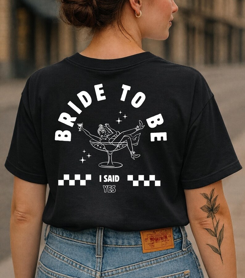 Puede incluir: Camiseta negra con un gr&aacute;fico blanco que dice "BRIDE TO BE" en arco. El gr&aacute;fico presenta a una mujer en una copa de champ&aacute;n, con el texto "I SAID YES" debajo. La camiseta la lleva una persona con vaqueros azules.
