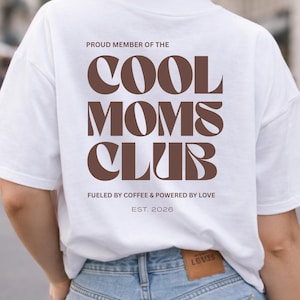 Könnte beinhalten: Weißes T-Shirt mit dem braunen Schriftzug "COOL MOMS CLUB", den Sätzen "PROUD MEMBER OF THE", "FUELED BY COFFEE & POWERED BY LOVE" und "EST. 2026". Das Shirt wird mit einer blauen Jeans getragen.