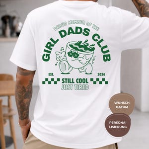 May include: White t-shirt with a green graphic on the back that reads "GIRL DADS CLUB" with a cartoon coffee cup character. The text "STILL COOL JUST TIRED" is below the graphic. The shirt also has the text "EST. 2026".