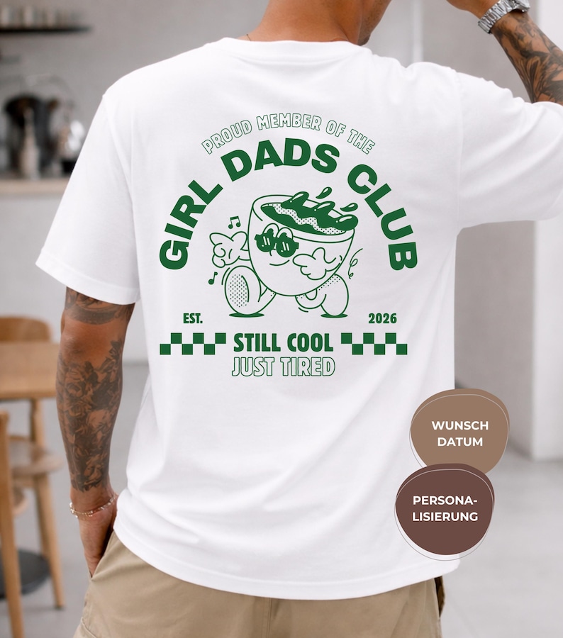 May include: White t-shirt with a green graphic on the back that reads "GIRL DADS CLUB" with a cartoon coffee cup character. The text "STILL COOL JUST TIRED" is below the graphic. The shirt also has the text "EST. 2026".