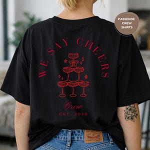 May include: Black t-shirt featuring red text and a champagne glass pyramid design. The text reads "WE SAY CHEERS" and "Crew EST. 2026". The shirt has a relaxed, oversized fit.