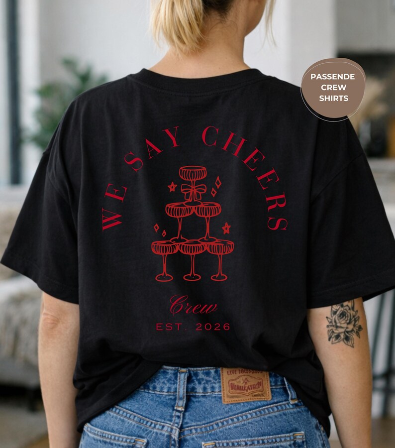 May include: Black t-shirt featuring red text and a champagne glass pyramid design. The text reads "WE SAY CHEERS" and "Crew EST. 2026". The shirt has a relaxed, oversized fit.